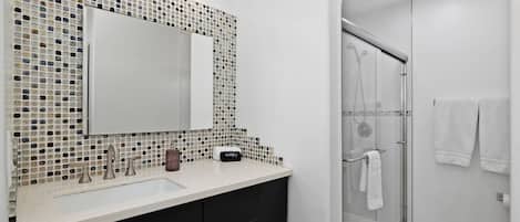 A modern bathroom with a mosaic tile backsplash, a large mirror, a glass shower enclosure, and a white sink with black cabinets.