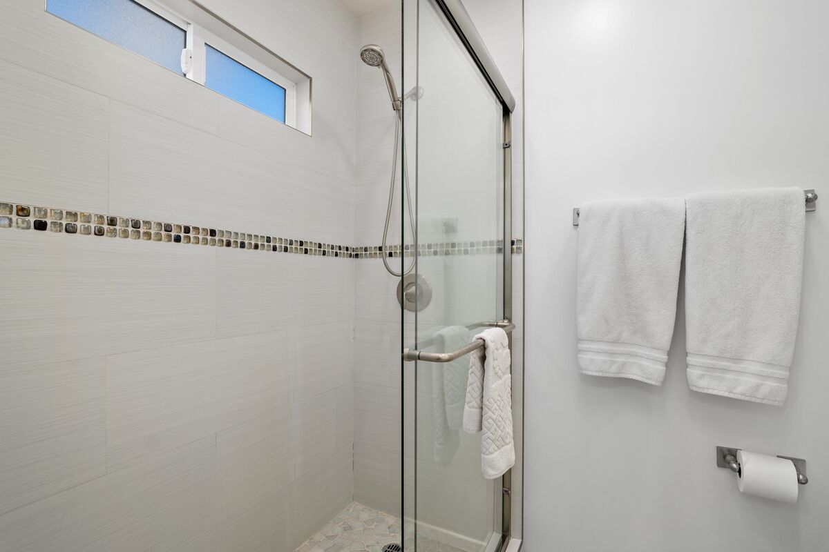 A glass-enclosed shower with a handheld showerhead, a towel rack with two towels, and a toilet paper holder on the wall.