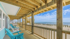 Une terrasse en bois avec des fauteuils Adirondack bleus, un escalier menant à une zone abritée, et une vue sur la plage et l’océan.