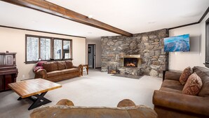 A spacious living room with a stone fireplace, leather sofas, a wooden coffee table, and a large window with a view of trees.