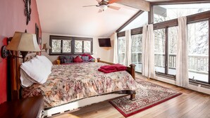 A bedroom with a large bed, a ceiling fan, a TV, and a view of the outdoors through large windows.