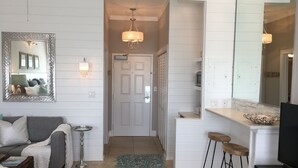 A modern interior with a grey sofa, a small round table, a blue rug, and a kitchen area with stools and a TV. - Spectacular Ocean View! Great Location On 30A   King Bed ,Newly Remodeled! (Santa Rosa Beach)