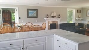 Amplia sala de estar con chimenea, comedor con sillas de madera y cocina con armarios blancos.