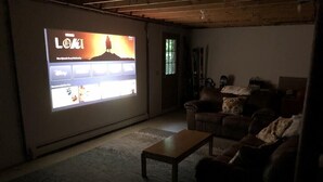 A dimly lit room with a large screen displaying a video game, a leather couch, a wooden coffee table, and a door with a window. - Cozy Mountain Escape to Southern Vermont (Wilmington)