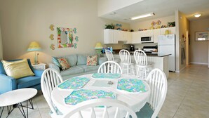 A living room with a blue sofa, a dining table with white chairs, and a kitchen in the background. - Caribbean Dunes 2BR/2BA with The Best Gulf View (Destin)