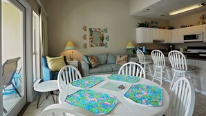 A dining area with white chairs and tables, a sofa, and a kitchen in the background. - Caribbean Dunes 2BR/2BA with The Best Gulf View (Destin)