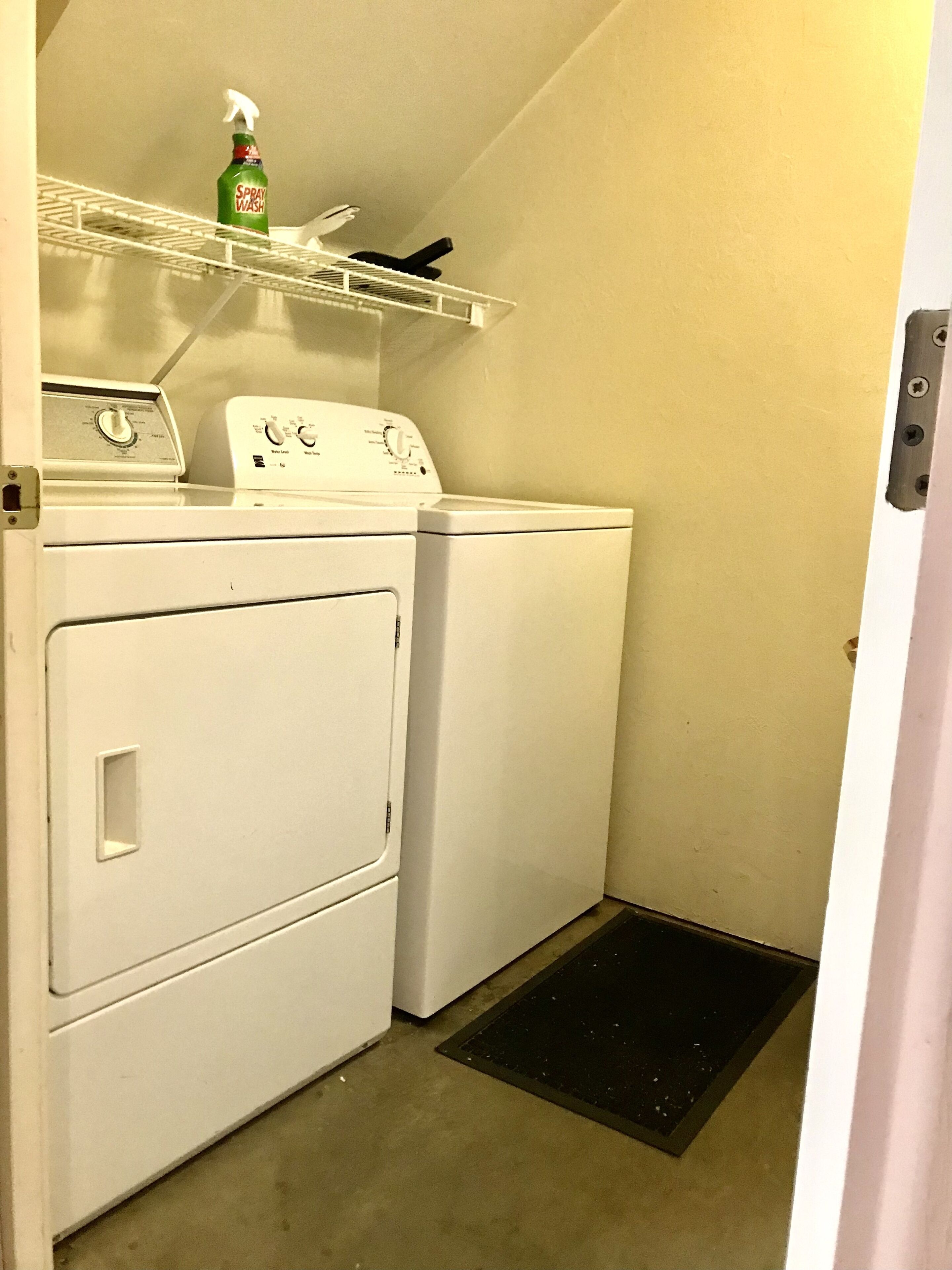 A laundry area with a front-loading washer and dryer, a shelf with a spray bottle, and a small vent.