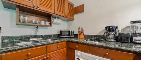 A kitchen with wooden cabinets, a granite countertop, a stainless steel sink, a microwave, a toaster, a coffee maker, and a blender.