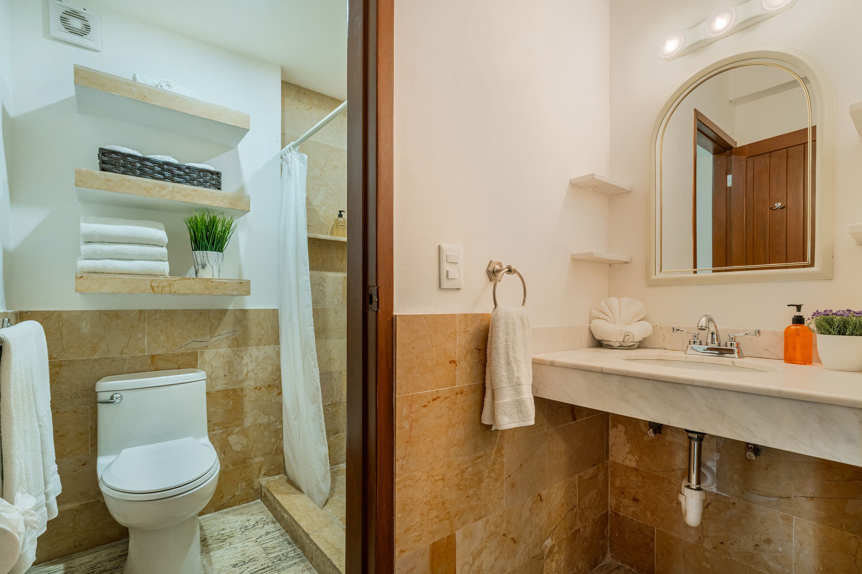 A bathroom with a toilet, shower, and sink. There are shelves above the toilet and a mirror above the sink.