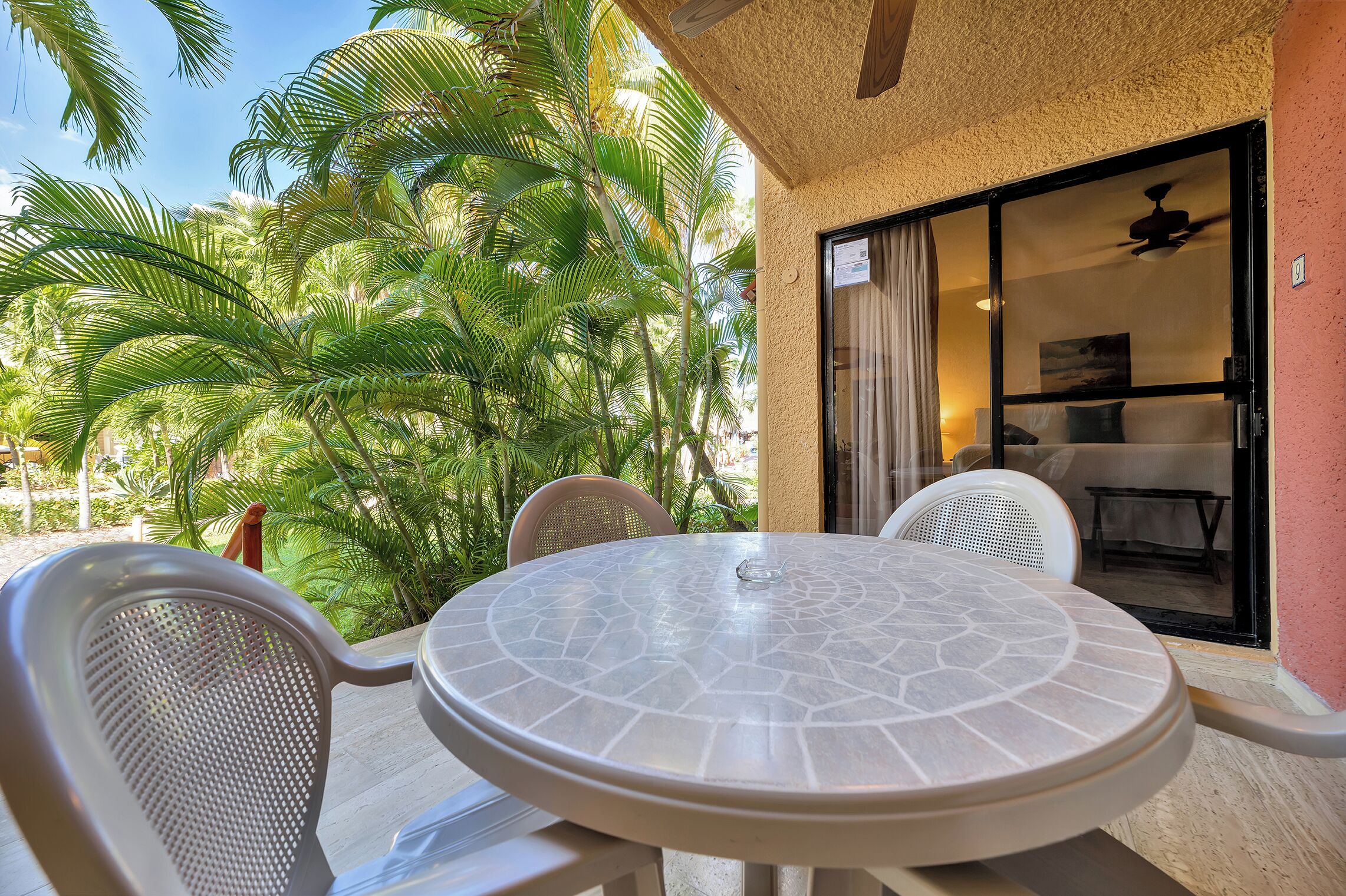 A round table with chairs, a ceiling fan, and a view of palm trees.