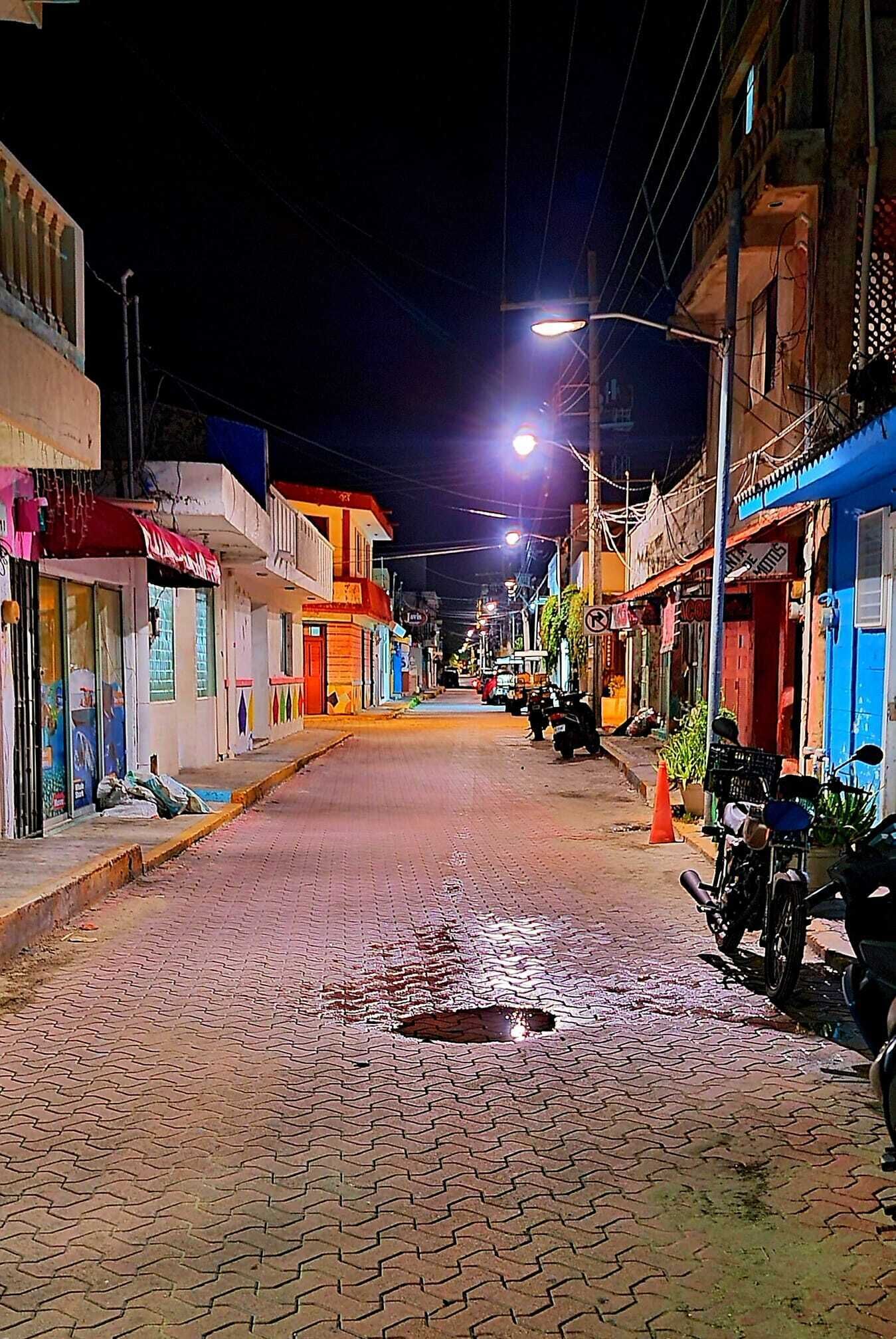 Cena noturna de rua com edifícios em ambos os lados, um caminho pavimentado e algumas motocicletas estacionadas.