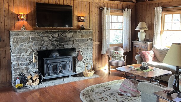 A rustic cabin interior with a stone fireplace, wooden beams, a flat-screen TV, and a cozy seating area with a table and chairs.