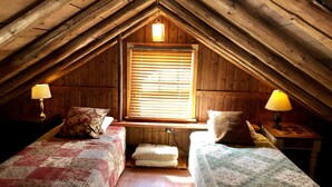 A cozy attic bedroom with two beds, wooden walls, a window with blinds, and two bedside lamps.