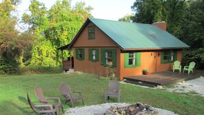 A cabin with a green roof and wooden siding, a fire pit in the front, and several Adirondack chairs.