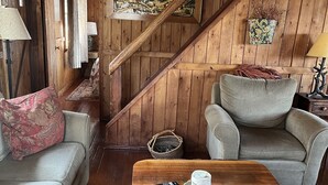 A cozy room with a wooden ceiling, a sofa, a chair, and a coffee table with a remote control and a candle.