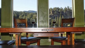 A wooden table with two chairs and two wine glasses, set against a backdrop of a forest and mountains through large windows. - Gorgeous riverfront, 4-BR (2-master suite) home! (Southfork)