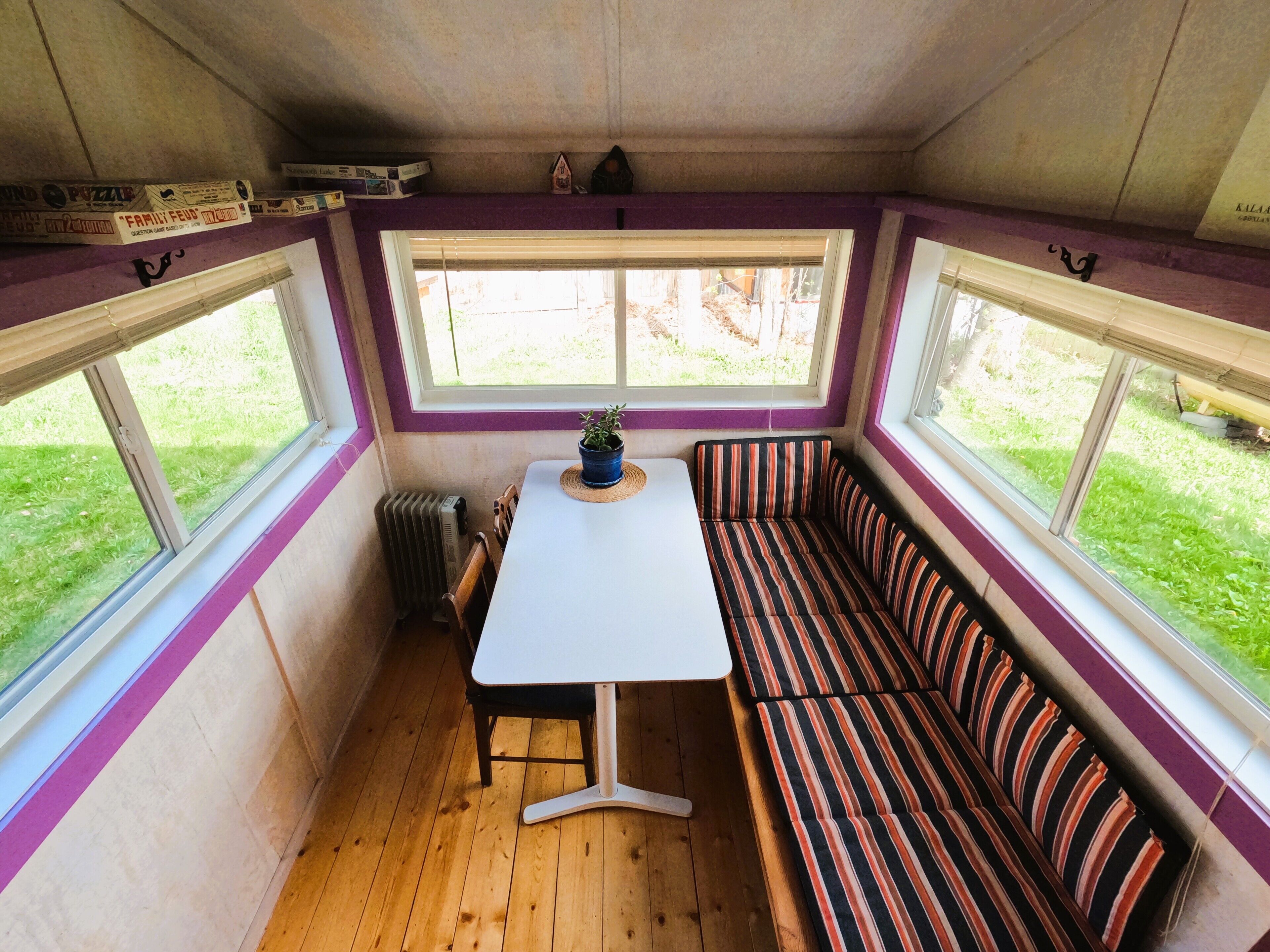 Habitació senzilla amb terra de fusta, taula blanca, una cadira de banc a ratlles i dues finestres que donen a una zona verda.