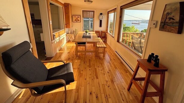 A spacious room with a wooden floor, a dining table, and a large window offering a view of the sea.