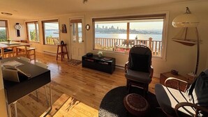 A spacious living room with a large window overlooking a body of water and a city skyline. There is a dining area with a table and chairs, a black entertainment unit, and a comfortable seating area with a chair and ottoman.