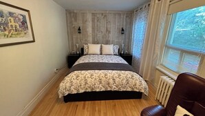 A bedroom with a wooden floor, a bed with a floral bedspread, two bedside lamps, a radiator, and a window with blinds.