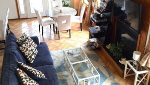 Ein geräumiges Wohnzimmer mit einem blauen Sofa, einem Couchtisch und einem Fernseher. Es gibt eine Essecke mit Tisch und Stühlen sowie eine Treppe, die zu einer oberen Ebene führt.