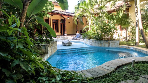 A swimming pool surrounded by lush greenery and stone walls, with a building in the background.