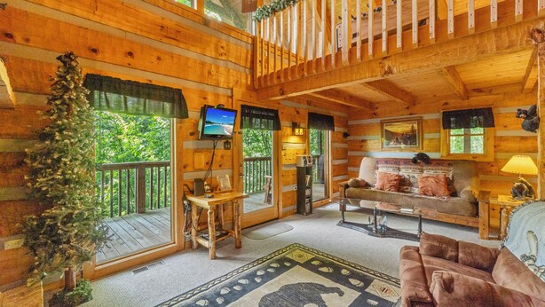 L’intérieur chaleureux d’un chalet, avec un décor en bois, une télévision, une cheminée et une grande fenêtre donnant sur un paysage verdoyant.