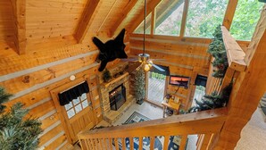 L’intérieur d’un chalet en bois, avec une cheminée, un ventilateur de plafond et une vue sur l’extérieur grâce à de grandes fenêtres.