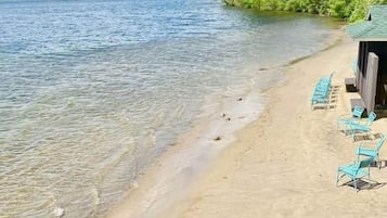 A lakeside view with a sandy beach, lounge chairs, and a forested area.