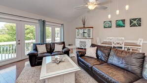 A spacious living room with leather sofas, a glass coffee table, and a ceiling fan. There are large windows and a view of greenery outside. - Siesta Key private beach access (Sarasota)