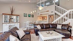 A spacious living room with a brown leather sectional sofa, a glass coffee table, and a white cabinet. There is a staircase leading to an upper level, and a dining area with white chairs. - Siesta Key private beach access (Sarasota)