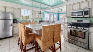 Une cuisine avec un plafond de couleur turquoise, des appareils électroménagers en acier inoxydable et des chaises en osier disposées autour d’un îlot central.