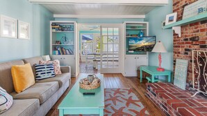 Un salon chaleureux avec un canapé, une table basse et un mur en briques. On y trouve des étagères remplies de livres et d’objets décoratifs, et une porte vitrée offre une vue sur l’extérieur.