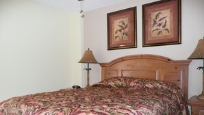 Ein Schlafzimmer mit einem hölzernen Kopfteil, einem blumigen Bettbezug, zwei gerahmten Pflanzenmotiven an der Wand und zwei Nachttischlampen.