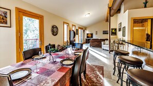 Un espace repas avec une longue table dressée pour le repas, une cuisine en arrière-plan et un coin bar avec des tabourets.