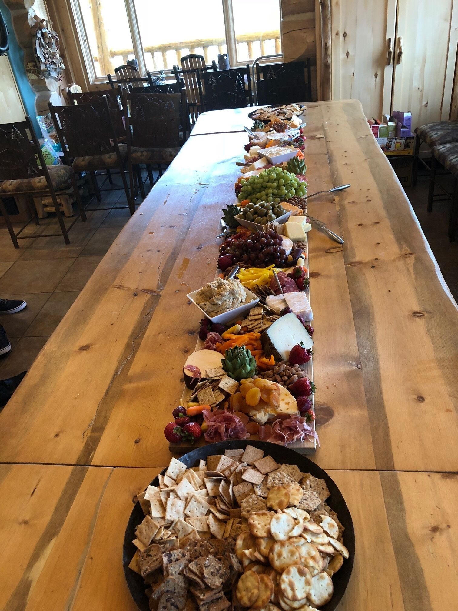 A long wooden table with a variety of cheeses, fruits, and crackers arranged for a buffet.