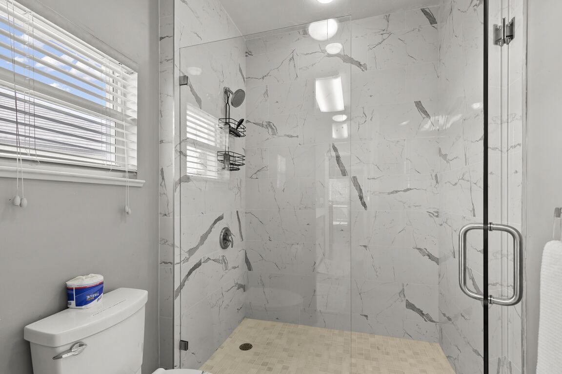 A modern bathroom with a glass shower enclosure, marble walls, and a toilet with a visible toilet paper roll.
