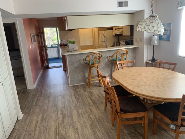 A dining area with a round wooden table and four chairs, a kitchen with white cabinets and a built-in oven, and a hallway leading to a bedroom.
