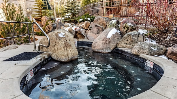 Un bain à remous entouré de gros rochers et de plaques de neige, avec une rambarde en métal et un panneau indiquant qu'il est interdit de se baigner.