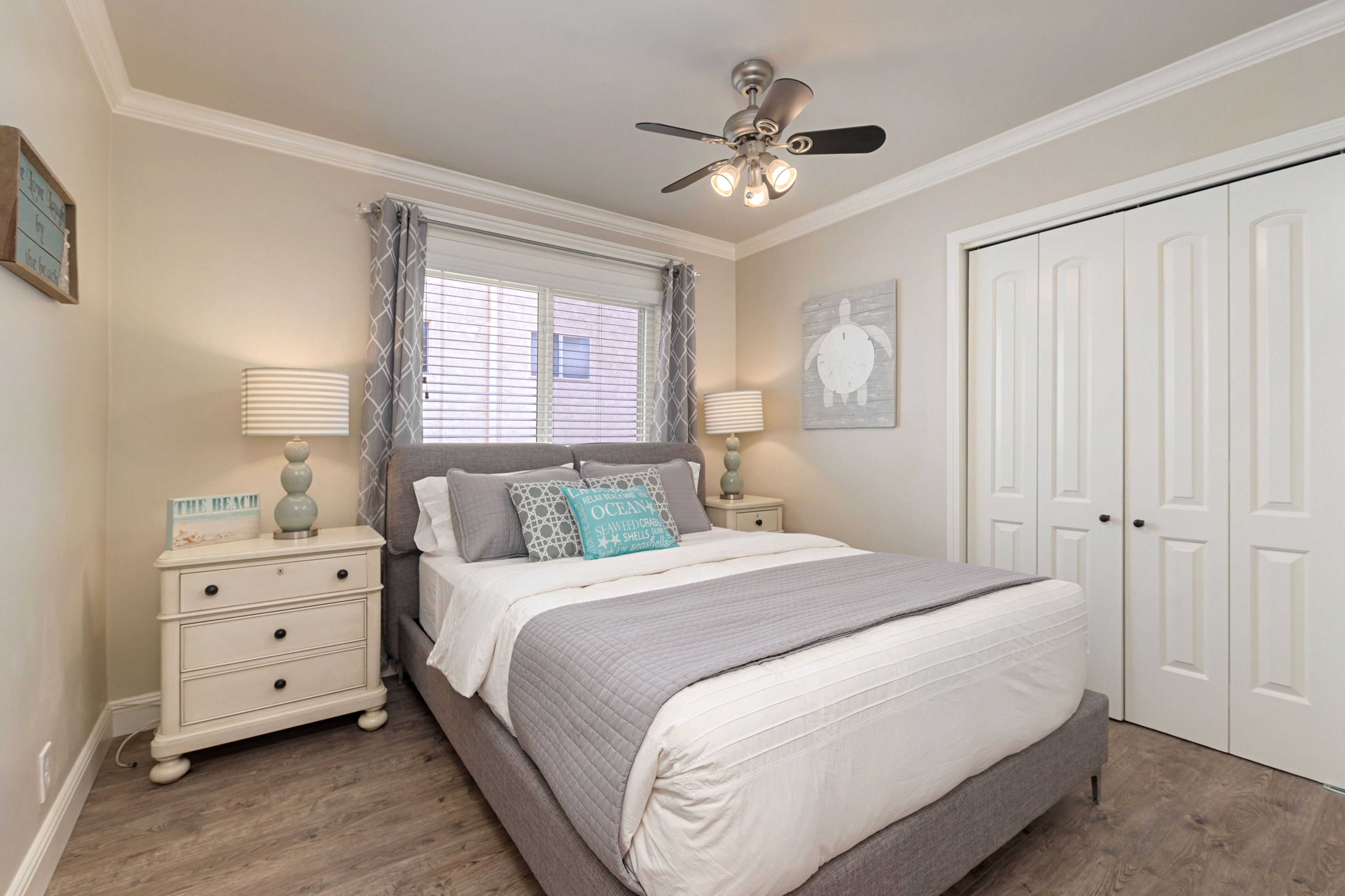 Une chambre à coucher comprenant un lit, des tables de chevet, un ventilateur de plafond, une fenêtre avec des rideaux, une porte et un mur orné d’un tableau.