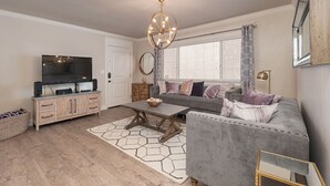 Ein Wohnzimmer mit grauem Sofa, einem Couchtisch, einem Fernseher auf einer Holzkommode und einer Deckenleuchte.
