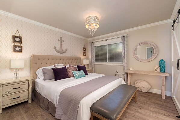 A bedroom with a large bed, a bedside table, a bench, a dresser, a chandelier, a window with curtains, and wall decorations including an anchor and framed pictures.