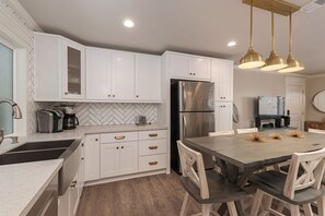 A modern kitchen with white cabinets, a stainless steel refrigerator, a wooden dining table, and pendant lighting.