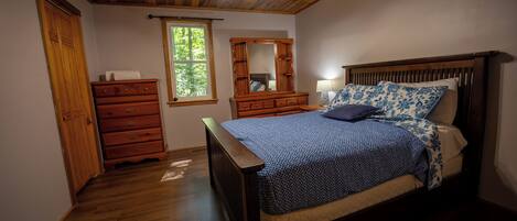 A bedroom with a wooden ceiling, a bed with a patterned bedspread, a dresser, a mirror, and a window.