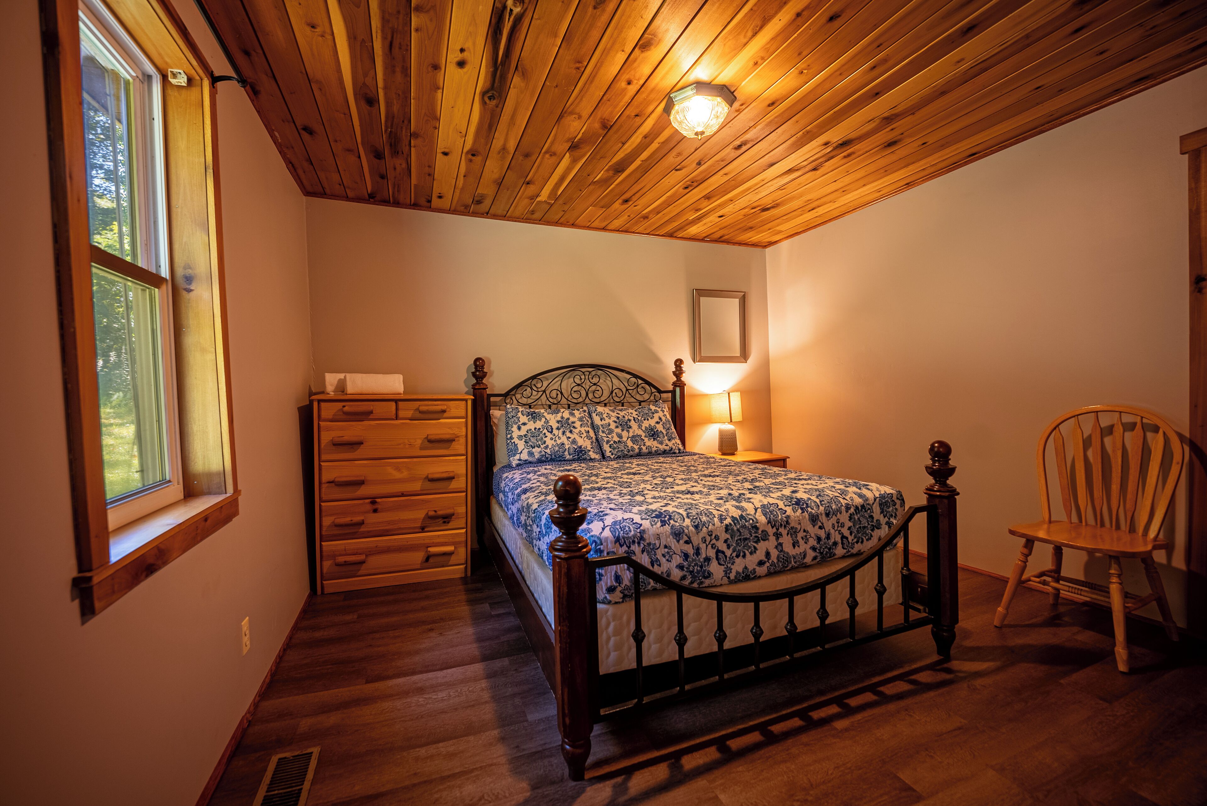 Un dormitorio con techo de madera, cama con estructura metálica, una silla de madera, una mesita de noche con una lámpara y una ventana con vista a árboles.