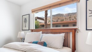 Un dormitorio con cabecera de madera, un ventanal con vistas a montañas y una mesita de noche con lámpara.