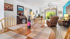 Un comedor con una mesa de madera, sillas y una botella de vino. Hay cuadros en las paredes y vistas al exterior a través de amplias ventanas.