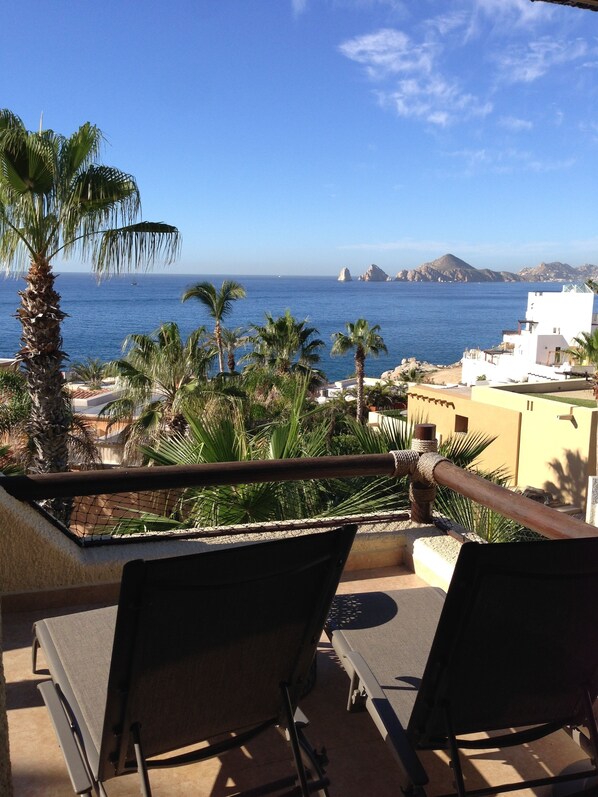 A balcony with two chairs overlooking a coastal view, palm trees, and a clear blue sky.