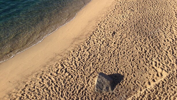 Ein Sandstrand mit klarem, blauem Wasser, Felsformationen und Gebäuden an einem Hügelhang.