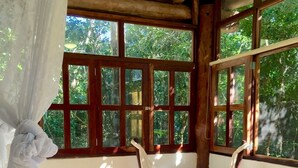 Una habitación con una silla hamaca, una cama y ventanas amplias con vista a la vegetación.